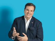 Mann in Anzug vor blauem Hintergrund, lächelnd und gestikulierend.