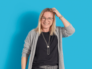 Lächelnde Person in Brille und grauem Cardigan vor blauem Hintergrund.