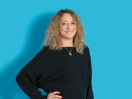 Person mit lockigem Haar in schwarzem Pullover vor blauem Hintergrund.