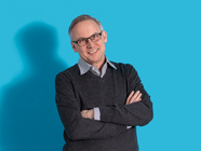 Älterer Mann mit Brille vor blauem Hintergrund, Arme verschränkt.