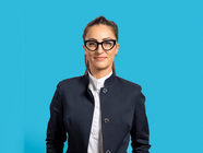 Frau mit Brille in schwarzer Jacke vor blauem Hintergrund.