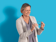 Person in einem hellen Blazer hält eine Brille vor blauem Hintergrund.