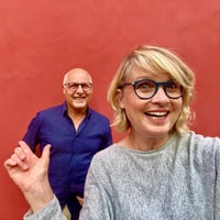 Frau mit blonder Haaren und blauer Brille winkt lächelnd, dahinter ein Mann im blauen Hemd, beide vor roter Wand.