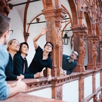 Fünf Menschen stehen auf einem verzierten Balkon und schauen lächelnd nach oben.