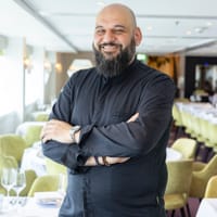 Mann mit Bart in schwarzem Hemd steht in elegantem Restaurant.
