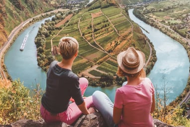 Zwei Personen sitzen auf Klippe mit Blick auf Fluss und Landschaft.