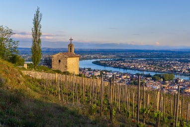 Kapelle auf einem Hügel mit Weinreben und Flussblick bei Sonnenuntergang.
