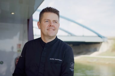 Person in dunkler Uniform lächelt an Bord eines Schiffes mit einer Brücke im Hintergrund.
