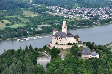 majestaetisch-ueber-dem-rhein-thronend-die-marksburg.jpg Burg am Fluss mit vorbeifahrendem Schiff und Stadt im Hintergrund.