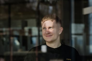 Person in schwarzer Kleidung steht hinter einer Glasscheibe, umgeben von Innenraumeinrichtung.