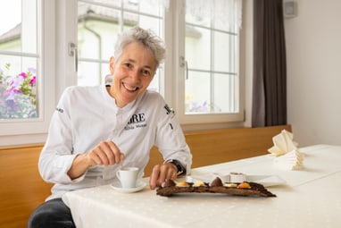 Frau in weisser Kochjacke sitzt am Tisch mit Kaffee und Dessert.
