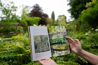 Person hält ein offenes Buch mit Gartenbildern vor üppiger grüner Landschaft.