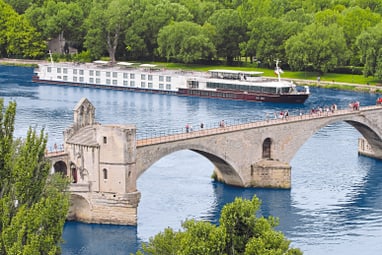 Alte Steinbrücke über Fluss mit Kreuzfahrtschiff und Bäumen im Hintergrund.