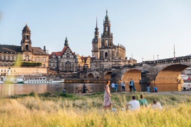 Personen entspannen im Gras mit Blick auf historische Gebäude und eine Brücke am Flussufer.