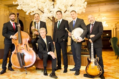 Sechs Männer in Anzügen mit Musikinstrumenten in einem eleganten Raum.