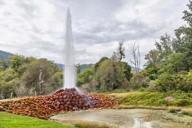 Wasserfontäne, die aus einem Steinhaufen in einer grünen Landschaft aufsteigt.