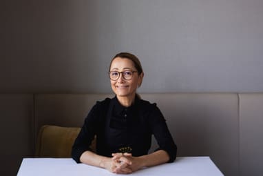 Frau mit Brille und dunklem Oberteil sitzt an einem Tisch.