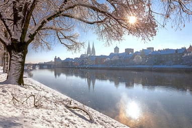 Verschneite Uferpromenade mit Bäumen und einer Stadt im Hintergrund, Sonne im Himmel.