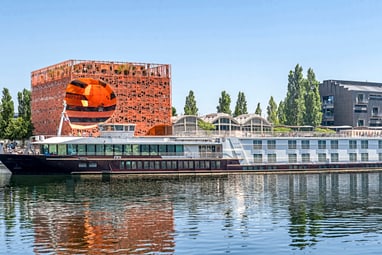 aufregend-modern-orange-cube-in-lyon.jpg Hausboot auf Fluss, moderne Gebäude im Hintergrund.