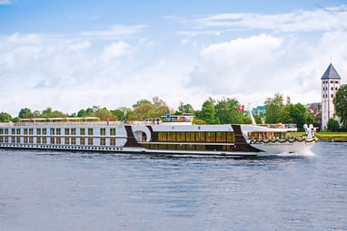 Grosses Flusskreuzfahrtschiff auf einem Fluss mit Bäumen und Turm im Hintergrund.