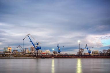 Flussufer mit Kränen und städtischer Skyline im Hintergrund unter bewölktem Himmel.