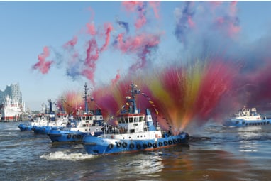 Mehrere Schlepperboote sprühen bunte Wasserfontänen im Hafen.