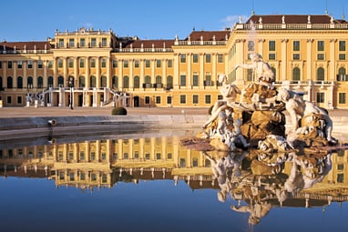 schloss-schoenbrunn-wien.jpg Historisches Gebäude mit gelber Fassade und einem Brunnen im Vordergrund, der sich im Wasser spiegelt.