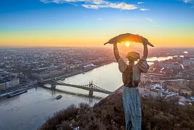 budapester-wahrzeichen-die-freiheitsstatue-auf-dem-gellertbe.jpg Monument über Budapest bei Sonnenuntergang mit Fluss und Brücke.