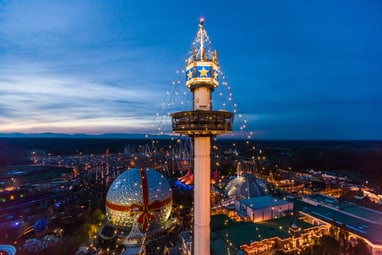 Abendansicht eines beleuchteten Vergnügungsparks mit Turm und Glaskuppel.