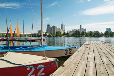 sommer-in-wien-blick-von-der-alten-donau-auf-die-stadtkuliss.jpg Hölzerner Steg am Wasser mit kleinen Segelbooten und Stadtansicht im Hintergrund.