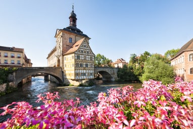 bamberg.jpg Historisches Gebäude auf einer Brücke über einem Fluss mit rosa Blumen im Vordergrund.