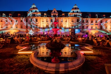 Beleuchtetes Schloss bei Nacht mit Springbrunnen im Vordergrund.