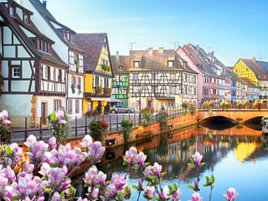 twerenbold_75876.jpg Malerische Fachwerkhäuser am Fluss mit Brücke und Blumen im Vordergrund.