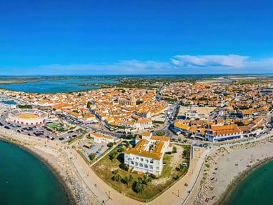 saintes-maries-de-la-mer-liegt-im-herzen-der-camargue-und-be.jpg Luftaufnahme einer Küstenstadt mit roten Dächern und Stränden.
