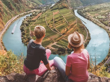 twerenbold_90174.jpg Zwei Personen sitzen auf Klippe mit Blick auf Fluss und Landschaft.