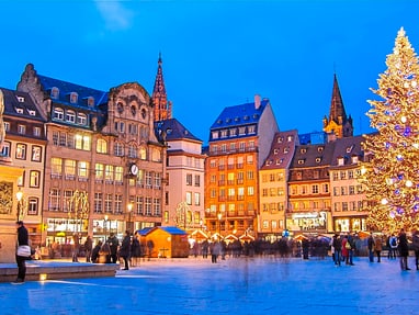 Weihnachtsbaum und Statuen auf beleuchtetem Platz bei Dämmerung.