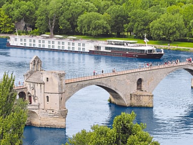 die-rhone-bei-der-pont-d-avignon.jpg Alte Steinbrücke über Fluss mit Kreuzfahrtschiff und Bäumen im Hintergrund.