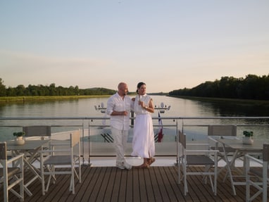 Zwei Personen in weisser Kleidung stehen auf dem Deck eines Bootes, umgeben von Tischen und Stühlen, mit Blick auf eine ruhige Wasserlandschaft.