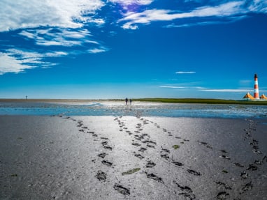 unesco-weltnaturerbe-wattenmeer.jpg Strandlandschaft mit Fussspuren im Sand, zwei Personen in der Ferne und einem Leuchtturm am Horizont.