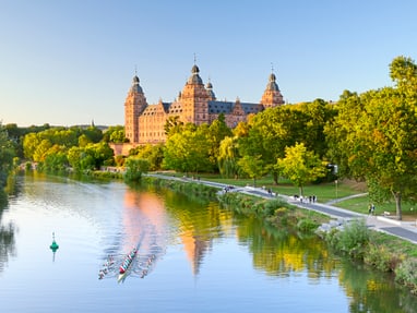 schloss-johannisburg-am-mainufer-in-aschaffenburg.jpg Grosses Schloss am Fluss mit Ruderboot, umgeben von Bäumen.