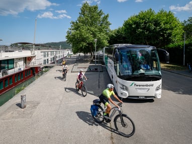 unterwegs-mit-schiff-bus-und-velo.jpg Menschen fahren mit Fahrrädern neben einem geparkten Reisebus und einem Flusskreuzfahrtschiff.