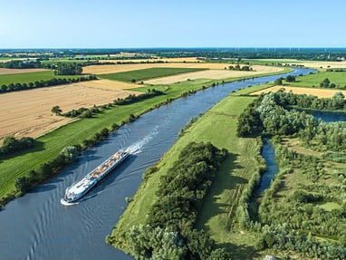 die-excellence-pearl-auf-der-weser.jpg Luftaufnahme eines Flusses mit einem Schiff und umliegenden Feldern.