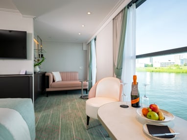 wohlfuehlorte-mit-blick-auf-den-fluss-die-suiten-und-kabinen.jpg Luxuriöses Zimmer mit Sofa, Tisch, Obstschale, Fenster mit Flussblick.