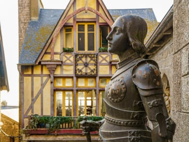 jeanne-de-arc-statue-in-der-normandie.jpg Statue eines Ritters vor einem historischen Gebäude.