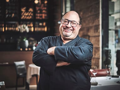 der-rhein-das-meer-und-antonios-cucina-9.jpg Ein lächelnder Koch in schwarzer Kleidung steht mit verschränkten Armen in einem Restaurant.