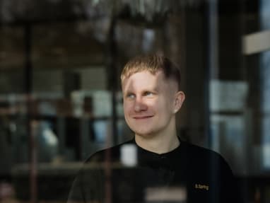 Person in schwarzer Kleidung steht hinter einer Glasscheibe, umgeben von Innenraumeinrichtung.