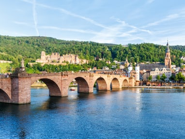 heidelberger-stadtensemble-neckarufer-altstadt-und-schlossru.jpg Steinbrücke über einen Fluss mit Stadt und Hügel im Hintergrund.