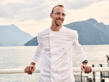 das-excellence-gourmetfestival-5.jpg Mann in weissem Kochkittel steht draussen vor einem See mit Bergen im Hintergrund.