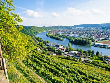 der-neckar-windet-sich-malerisch-durchs-tal.jpg Panorama einer Flusslandschaft mit Weinbergen und einer Stadt im Hintergrund.