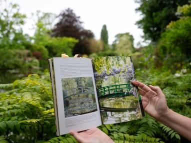 monets-seerosenteich-in-giverny.jpg Person hält ein offenes Buch mit Gartenbildern vor üppiger grüner Landschaft.